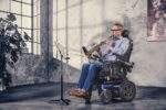 A man playing a clarinet while seated in an electric wheelchair, with sheet music on a stand.