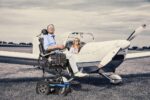Individual in a wheelchair with controls beside a light aircraft on a grassy field.