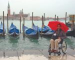 A man in a wheelchair holding a red umbrella beside gondolas in Venice, Italy.