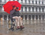 Individual in a wheelchair taking photographs with a camera while using a red umbrella.