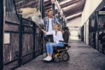 A woman in a wheelchair reaching out to a horse in a stable while a man stands nearby.