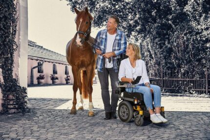 A man holding a horse's reins while a woman in a wheelchair sits beside him, both outdoors.