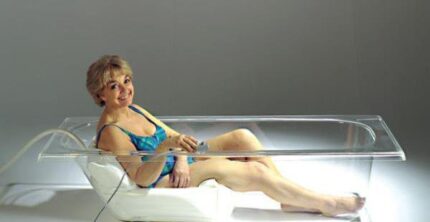 A woman sitting in a transparent bathtub, holding a device connected to it.