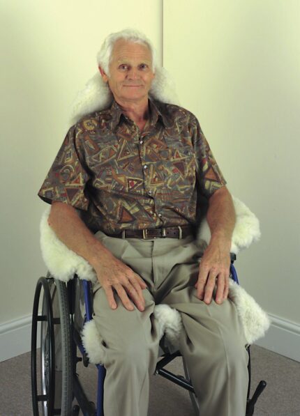 Older man seated in a wheelchair covered with a sheepskin throw, wearing a patterned shirt.