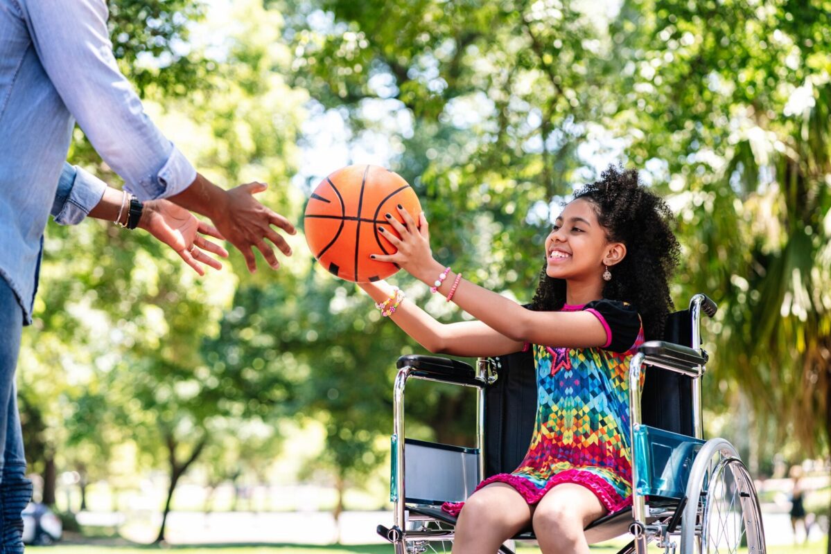 A young girl in a wheelchair reaching out with a basketball while smiling at a man.