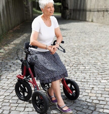 Image: A lady having a rest sitting on a red rollator in a cobbled street smiling using the Taima XC from Dietz Rehab