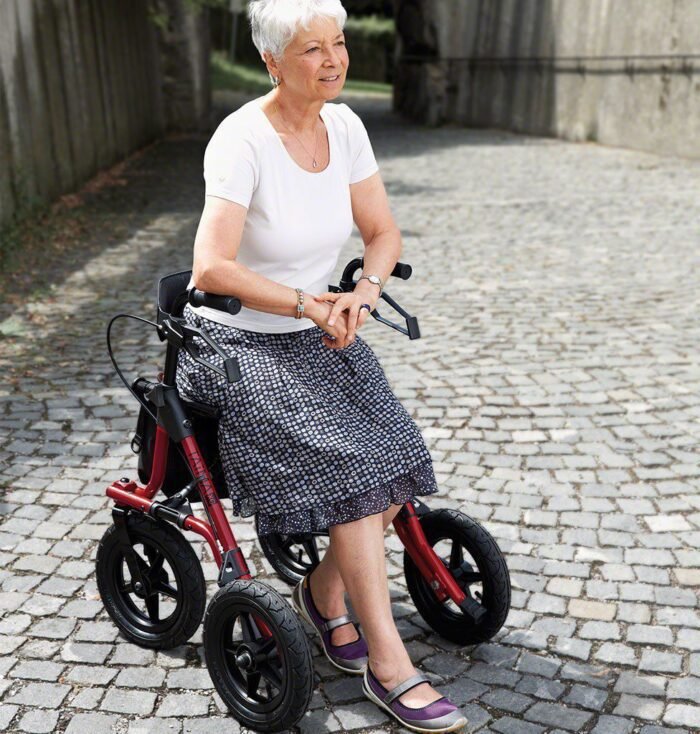 Image: A lady having a rest sitting on a red rollator in a cobbled street smiling using the Taima XC from Dietz Rehab