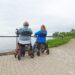 Two individuals seated in mobility walkers facing a lake with a paved path beside them.