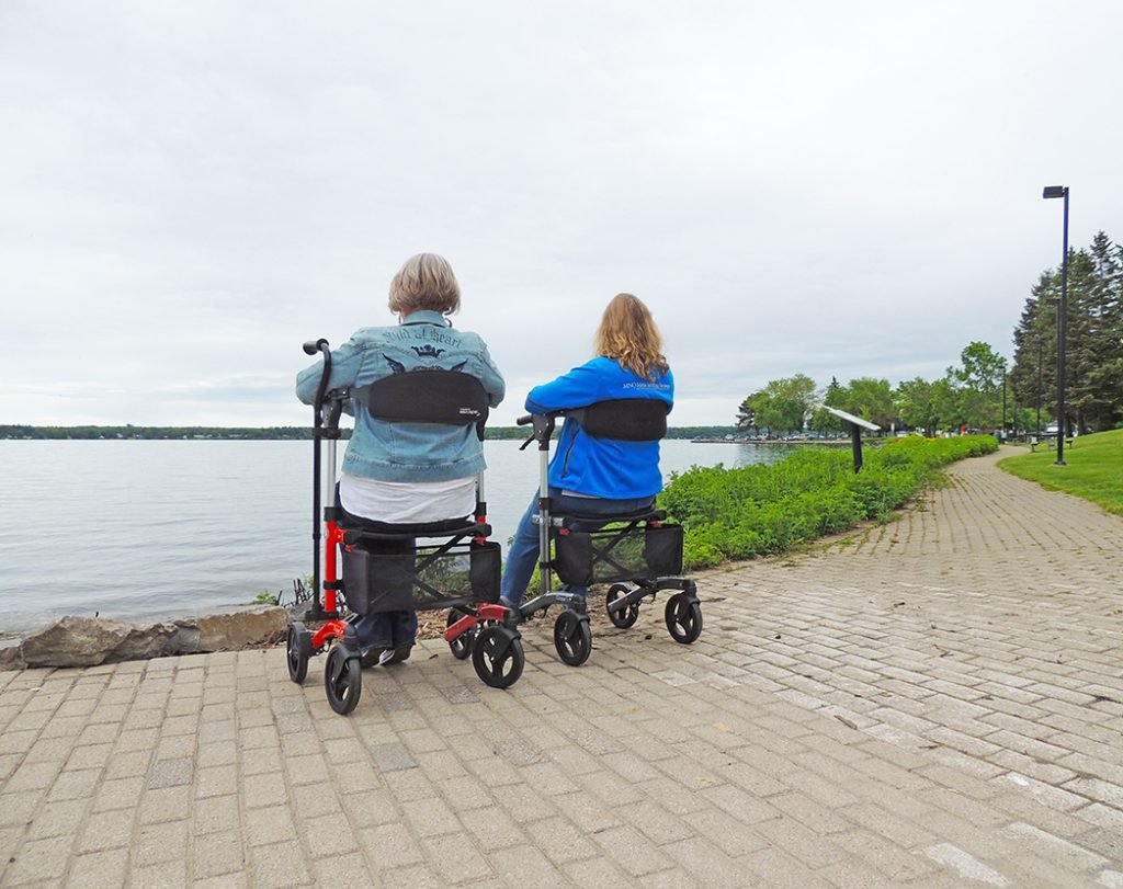 5 Essential Tips for Using a Rollator Safely and Comfortably 1 Younique Healthcare | Wheelchairs, Powerchairs, Ramps, Healthcare, Rehab, Mobility, Scooters in the West Midlands | 0121 630 3850 Two individuals seated in mobility walkers facing a lake with a paved path beside them.