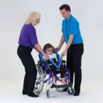 Two individuals assisting a child in a manual transfer sling attached to a wheelchair.