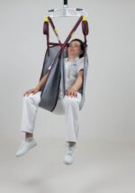 A person seated in a patient lift sling suspended from a ceiling hoist system.