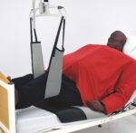 A patient model in a red garment being lifted with a leg support sling in a healthcare setting.
