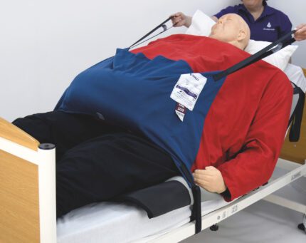 A training dummy dressed in a red and blue outfit, positioned on a hospital bed with support straps.