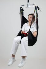 A woman seated in a black patient sling suspended from a lifting device.