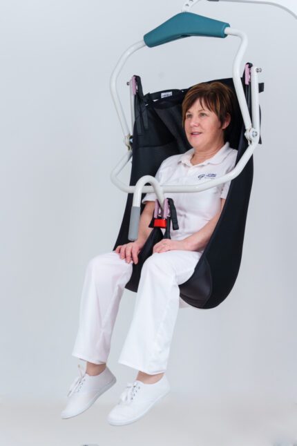 A person seated in a patient lifting sling, suspended from a ceiling track system.