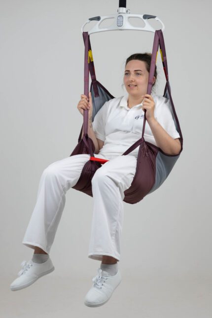 A person seated in a Silva Deluxe patient lifting sling, suspended from a ceiling lift system.