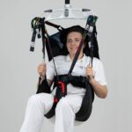 Individual seated in a Tinkham sling, suspended from a ceiling-mounted lift system.